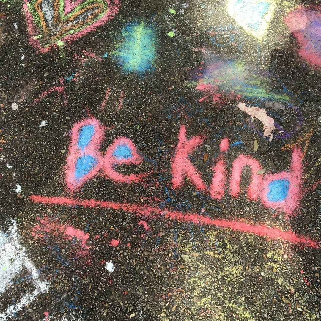 The words Be Kind spray painted in pink on the pavement. 